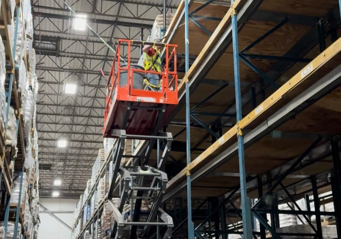 Technician on scissor lift dusting warehouse rafters